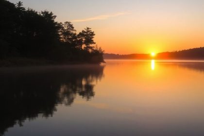 Sunrise over a calm lake with lush trees.